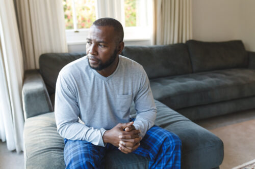 Worried, sad african american man thinking, sitting on couch looking out the window