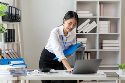Focused Businesswoman at Work: A cheerful and focused young businesswoman efficiently manages her workload, using a laptop and organizing files in a bright and modern office setting